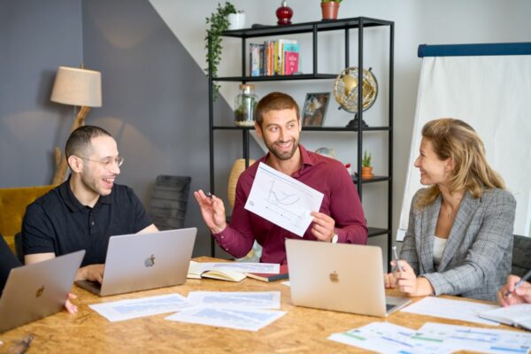 Three professionals engaging in a cheerful discussion around a wooden table, with laptops and documents in front of them. The person in the centre holds a paper with a graph, gesturing as if explaining a point, while the others listen attentively, with visible smiles.