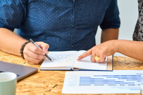 Close-up of two individuals collaborating over a notebook with hand-drawn flowcharts, with one person writing and the other pointing at the chart, on a table with a laptop and printed documents.