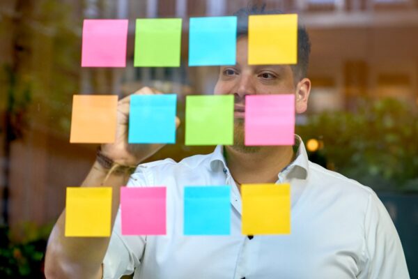 A man in a white shirt standing behind a glass pane covered with multicoloured sticky notes, appearing contemplative as he looks at the notes.