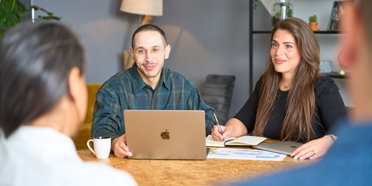 People Process Change-9214-min Team members at a table with laptops, a man smiling, engaging in discussion.
